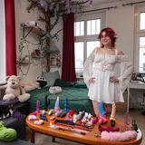 Junge Frau in weißem Kleid mit Elfenohren posiert in ihrer eigenen Wohnung vor ihrer Sammlung von Sexspielzeugen