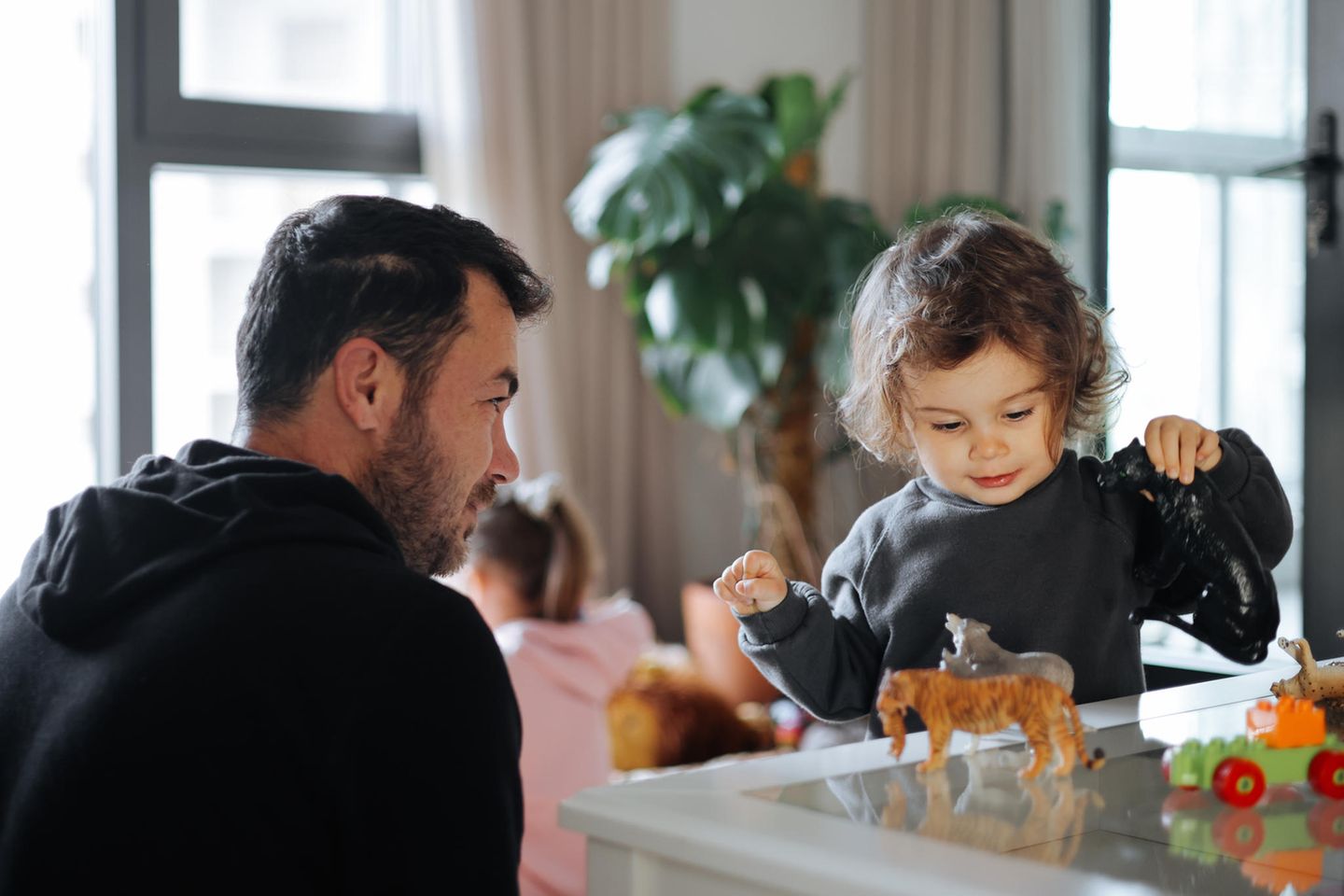 Tagesvater Ein Mann schaut zu wie ein Kind an einem Tisch mit Tierfiguren spielt. Ein anderes Kind spielt im Hintergrund.