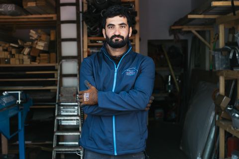 Lokman Eldemir steht mit verschränkten Armen, in die Kamera blickend vor der Werkstatt eines Dachdeckerbetriebes