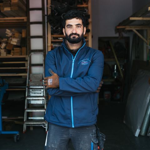 Lokman Eldemir steht mit verschränkten Armen, in die Kamera blickend vor der Werkstatt eines Dachdeckerbetriebes