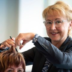 Eine Friseurin lacht in die Kamera und schneidet ihrer Kundin die Haare