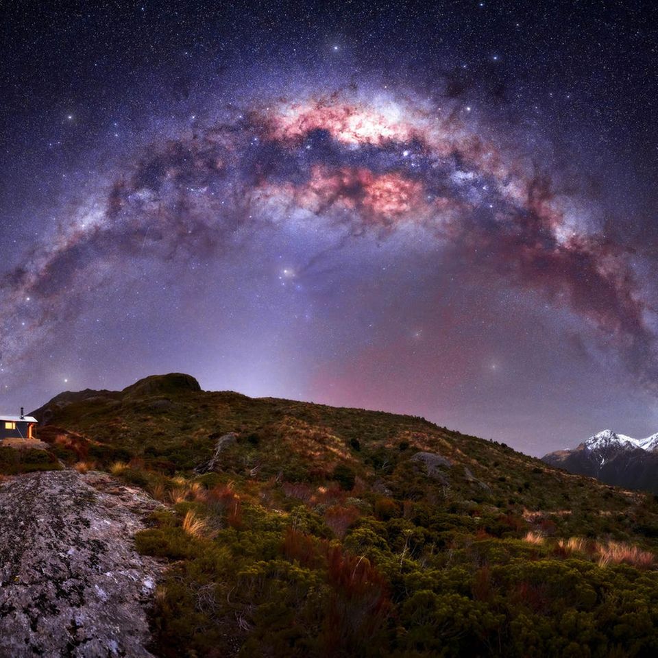 Eine violette Milchstraße über felsigem Gelände mit einer kleinen beleuchteten Hütte