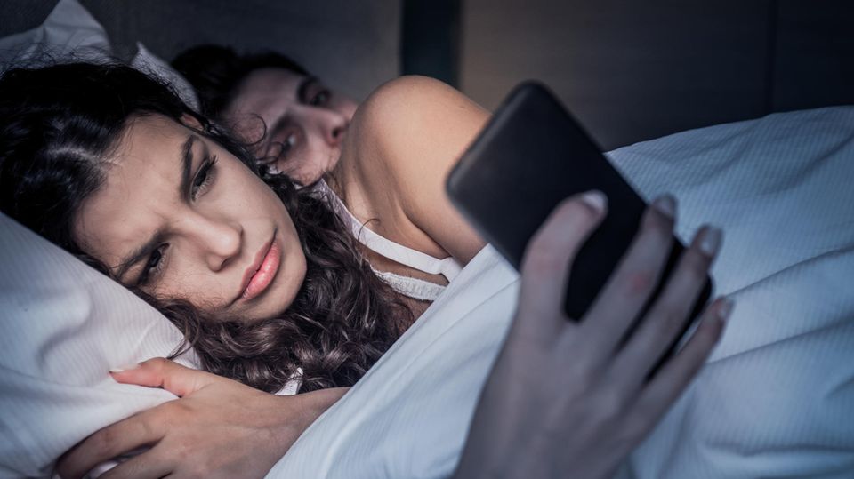 Paar liegt im Bett, sie sieht auf ihr Handy, er blickt misstrauisch hinüber zu ihr