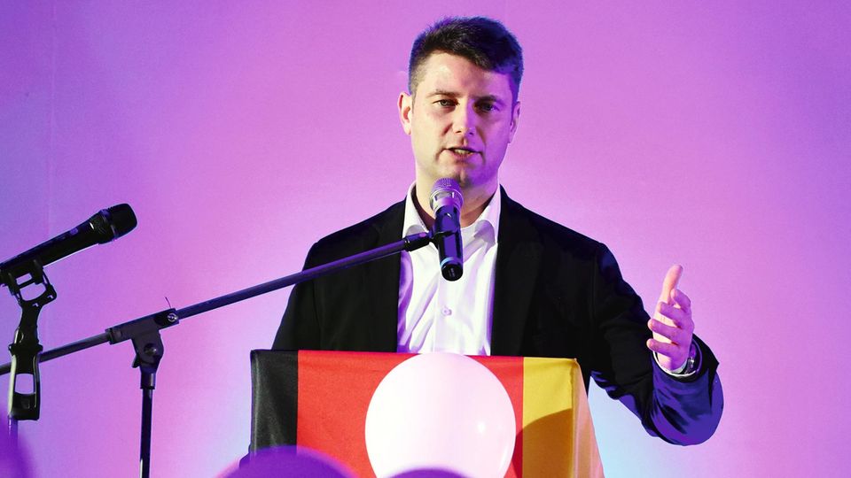 AfD-Politiker René Aust spricht auf einem Podium mit einer Deutschlandfahne in ein Mikrofon