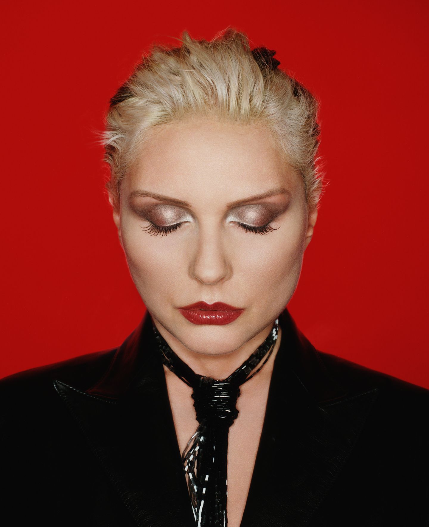 Debbie Harry vor rotem Hintergrund mit strenger Frisur und geschlossenen Augen