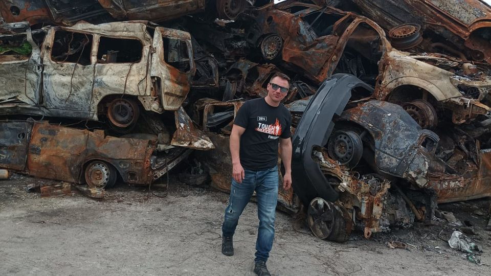Reiseführer Svet Moiseev vor den Trümmern von im Krieg zerstörter Autos bei Kiew