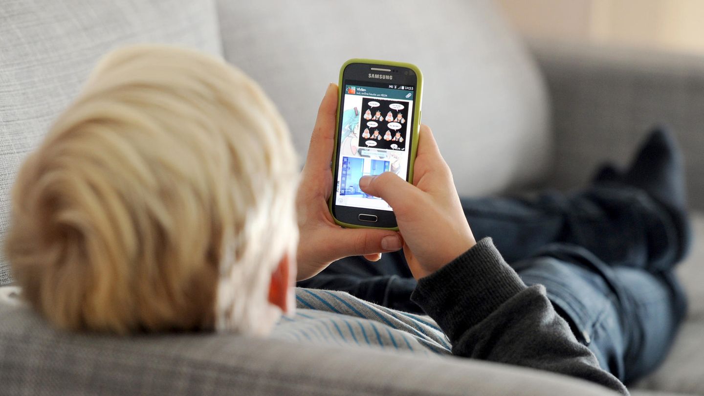 Ein blonder Junge liegt auf dem Sofa und blickt auf das Smartphone in seiner Hand