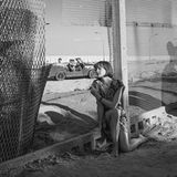 Eine juge Frau sitzt an einem geöffneten Maschendrahtzaun und schaut durch den Durchgang. Auf der anderen Seite steht ein Auto.