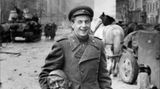 Jewgeni Dolmatowski hält einen Messingkopf in der Hand und lächelt. Im Hintergrund sind Pferde und Panzer zu sehen.