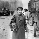 Jewgeni Dolmatowski hält einen Messingkopf in der Hand und lächelt. Im Hintergrund sind Pferde und Panzer zu sehen.