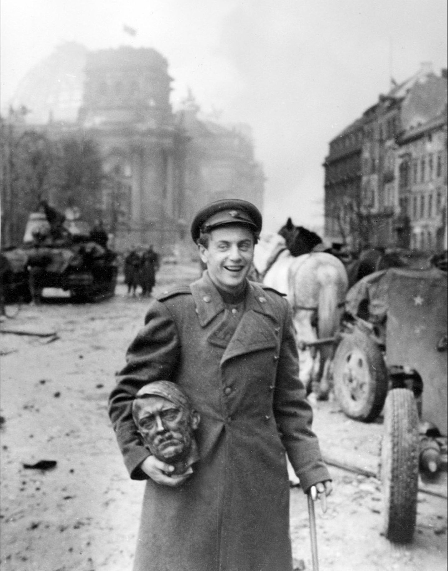 Jewgeni Dolmatowski hält einen Messingkopf in der Hand und lächelt. Im Hintergrund sind Pferde und Panzer zu sehen.