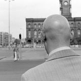 Ein mann mit Glatze wird nah von hinten fotografiert. Er steht an einer großen unbefahrenen Straße an einer Ampel.