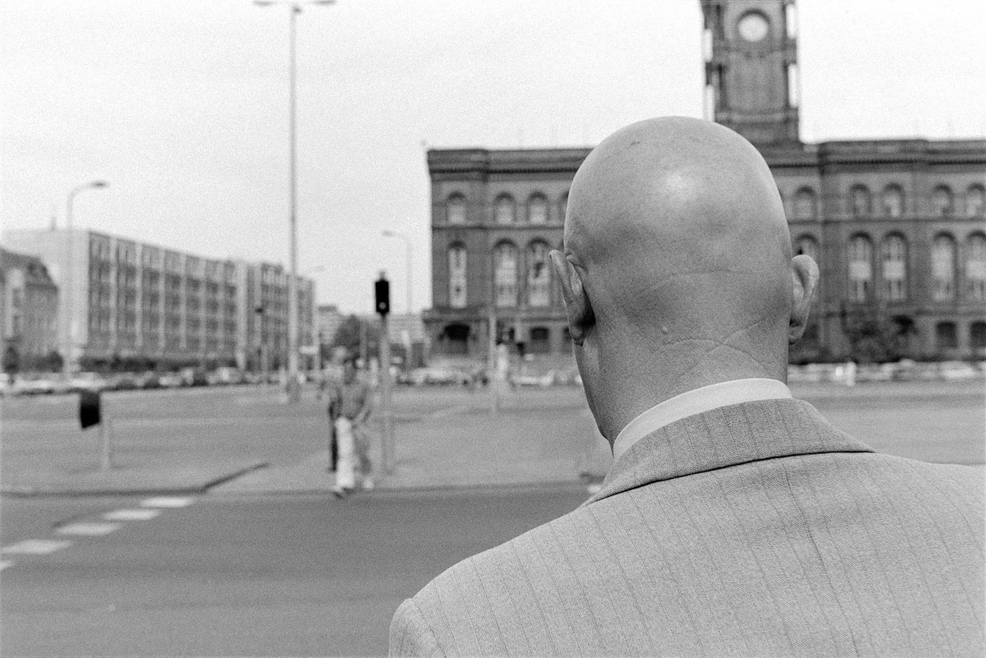Ein mann mit Glatze wird nah von hinten fotografiert. Er steht an einer großen unbefahrenen Straße an einer Ampel.