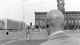 Ein mann mit Glatze wird nah von hinten fotografiert. Er steht an einer großen unbefahrenen Straße an einer Ampel.