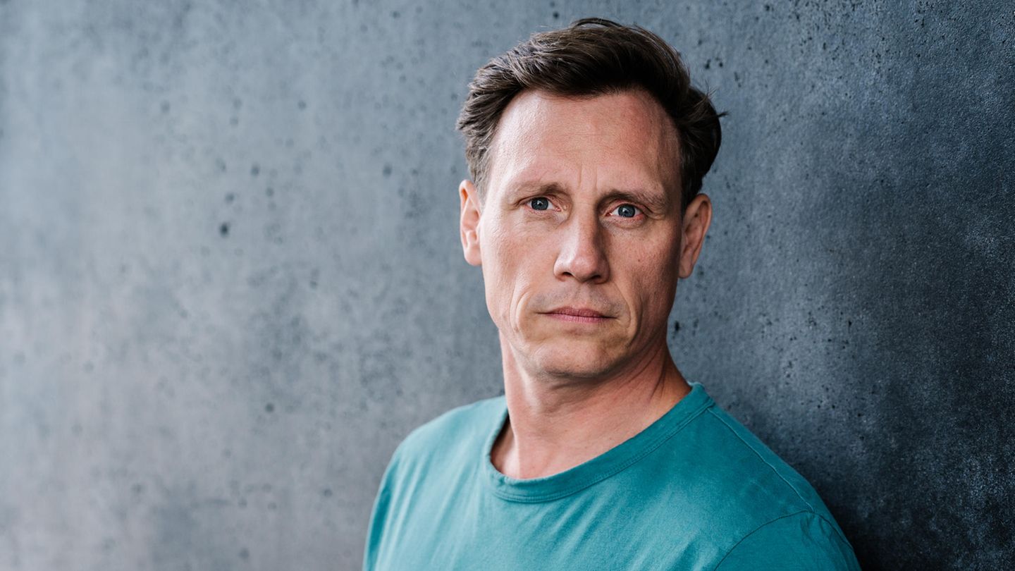 Schauspieler Patrick Kalupa in einem petrolfarbenen T-Shirt vor einer grauen Wand