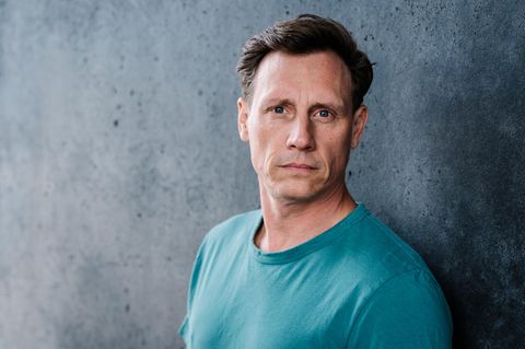 Schauspieler Patrick Kalupa in einem petrolfarbenen T-Shirt vor einer grauen Wand