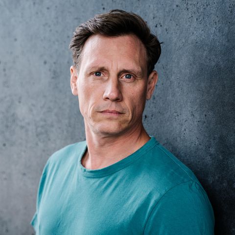 Schauspieler Patrick Kalupa in einem petrolfarbenen T-Shirt vor einer grauen Wand