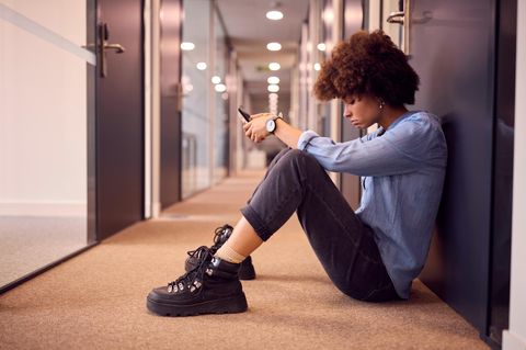 Eine junge Frau sitzt auf einem Flur in einem Büro auf dem Boden und schaut frustriert