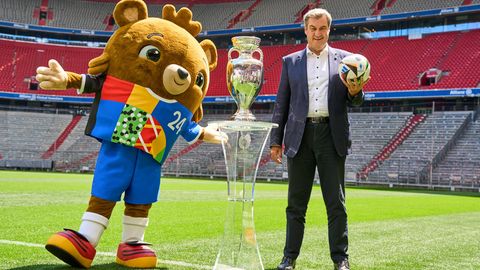 Markus Söder steht neben dem Fußball-EM Maskottchen und dem EM Pokal im Stadion
