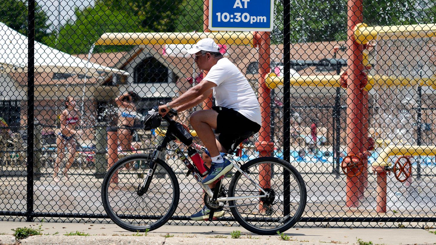 Ein Mann fährt bei heißem Wetter mit dem Fahrrad auf den Woodland Trails des River Trails Park District in Mount Prospect, Illinois. Im Hintergrund vergnügen sich Kinder lieber auf einem Wasserspielplatz