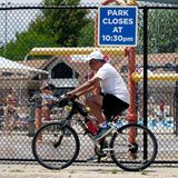 Ein Mann fährt bei heißem Wetter mit dem Fahrrad auf den Woodland Trails des River Trails Park District in Mount Prospect, Illinois. Im Hintergrund vergnügen sich Kinder lieber auf einem Wasserspielplatz