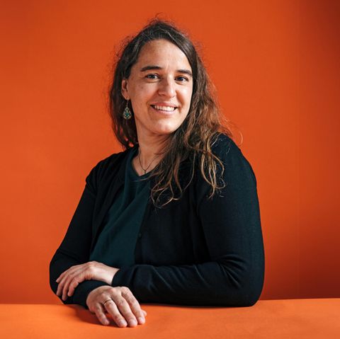Heike Heubach sitzt an einem Tisch und lächelt in die Kamera. Sie trägt die langen Haare offen und Ohrringe.