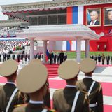 Die beiden Machthaber verfolgen das Spektakel im Schatten eines Unterstands mit antiker Anmutung. Im Hintergrund hängen übergroße Porträtbilder von Kim und Putin. Die Bildsprache spricht Bände