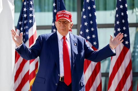 Donald Trump steht mit ausgebreiteten Armen vor vier Flaggen der USA