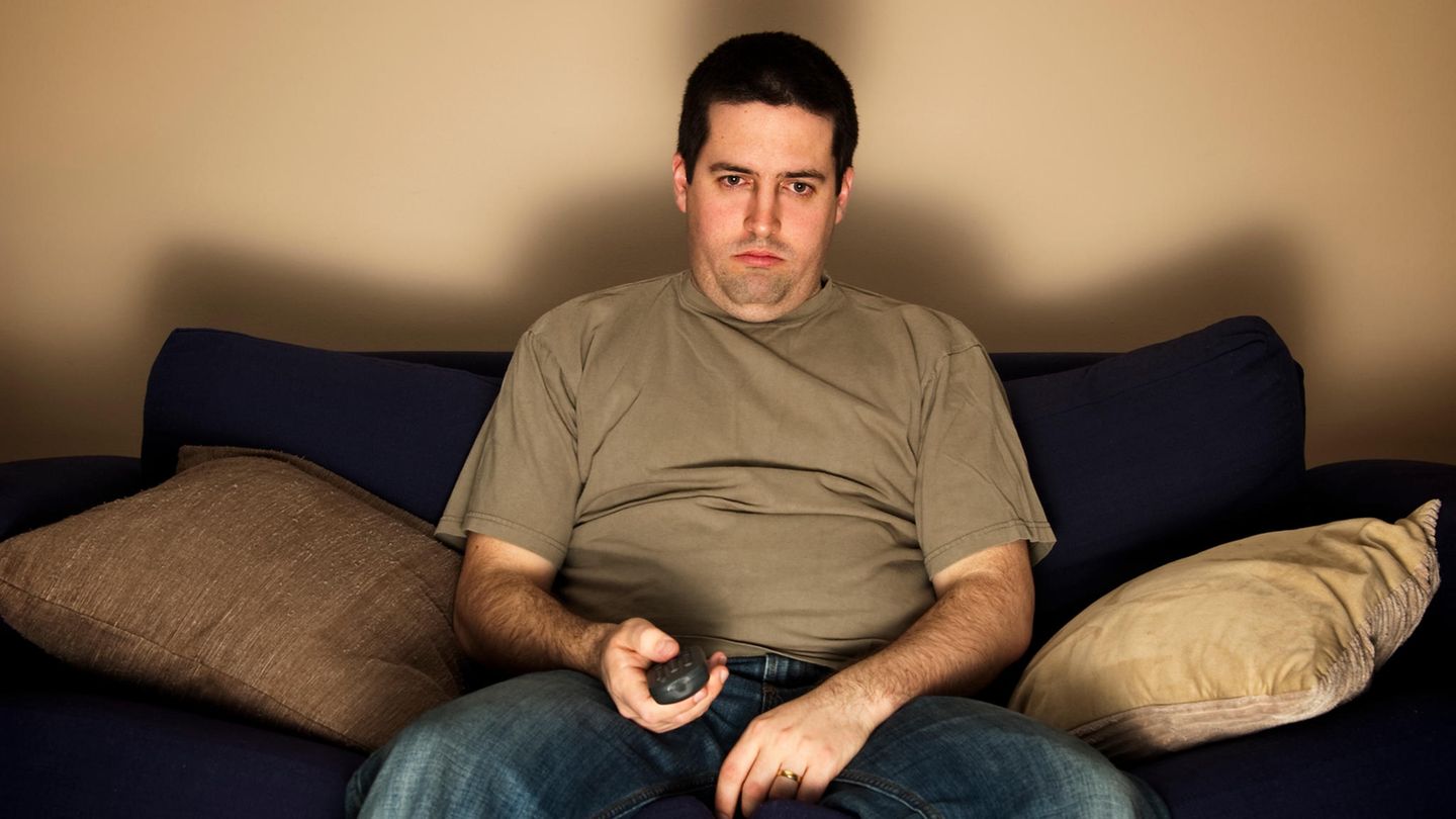 Mann sitzt lustlos auf einem Sofa mit Fernbedienung in der Hand