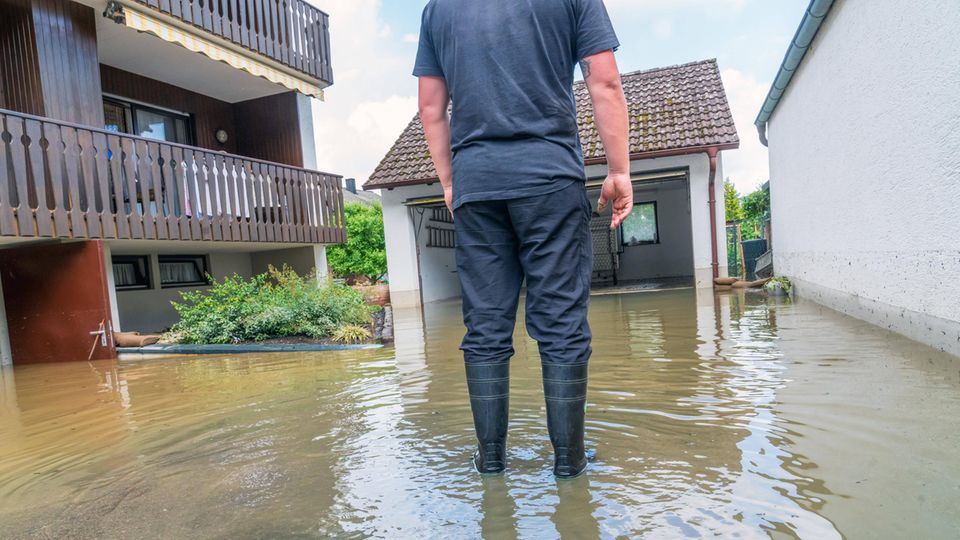 Ein Mann steht mit Gummistiefeln vor einem Haus und einer Garage, die unter Wasser stehen