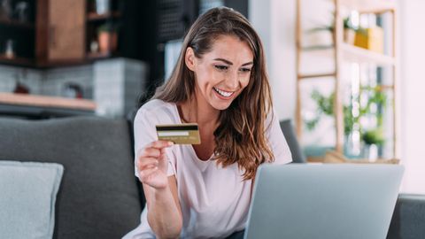 Amex Offers: Frau sitzt mit Kreditkarte in der Hand auf einem Sofa vor dem aufgeklappten Laptop.