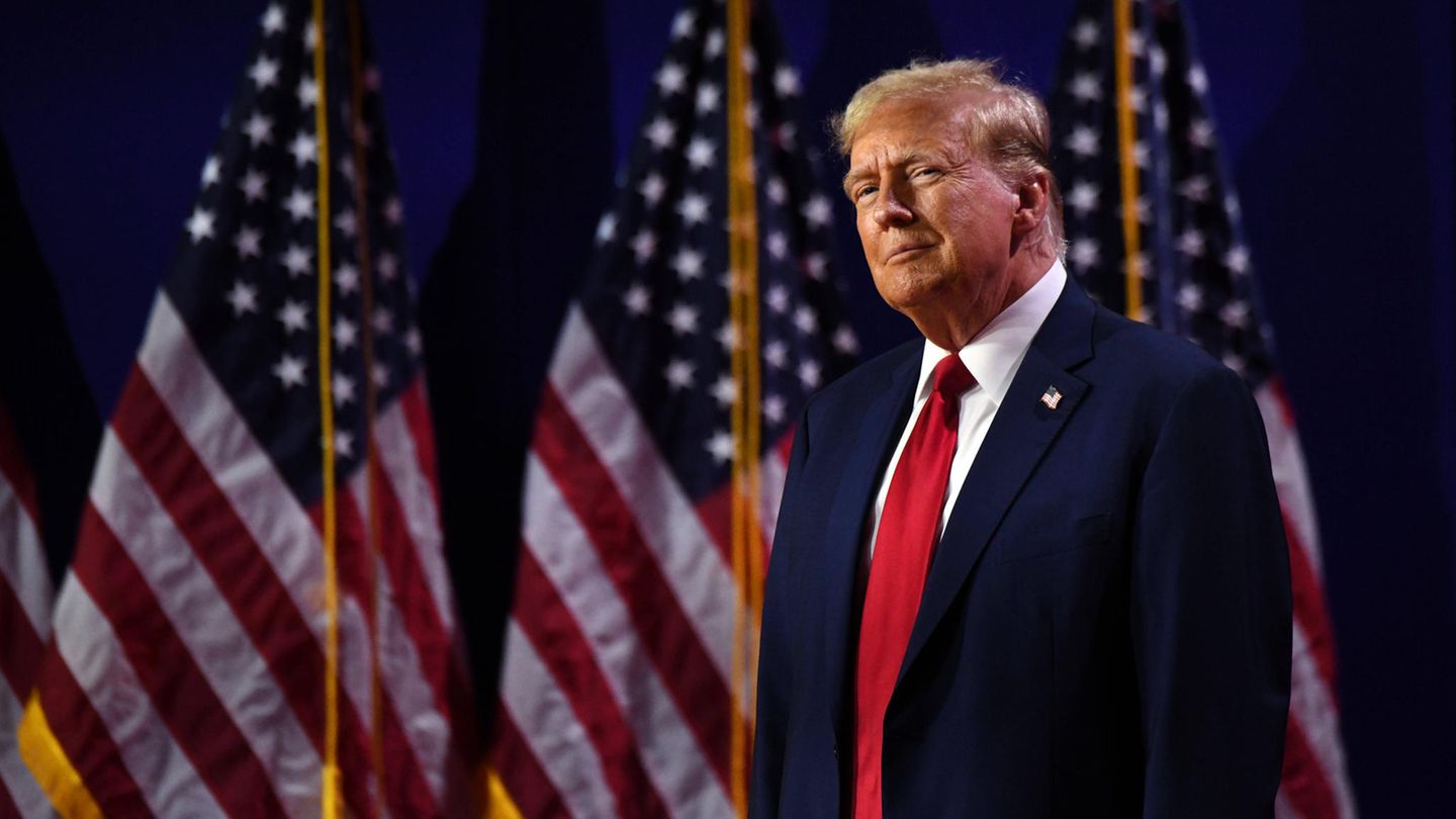Donald Trump steht vor amerikanischen Flaggen und sieht triumphierend aus