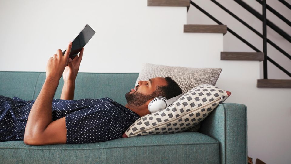 Mann liegt mit Kopfhörern und Tablet entspannt auf der Couch und schaut eine Serie