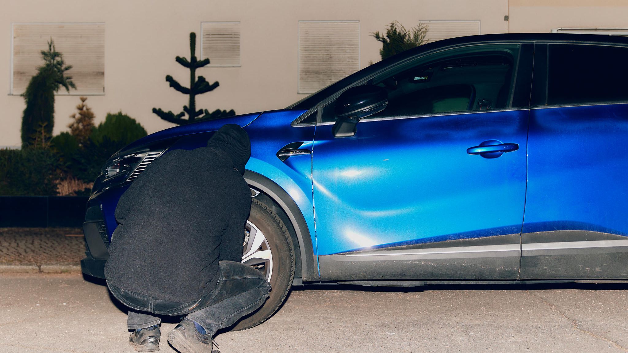 Dunkel gekleideter Mann macht sich nachts am linken Vorderrad eines blauen SUVs zu schaffen.