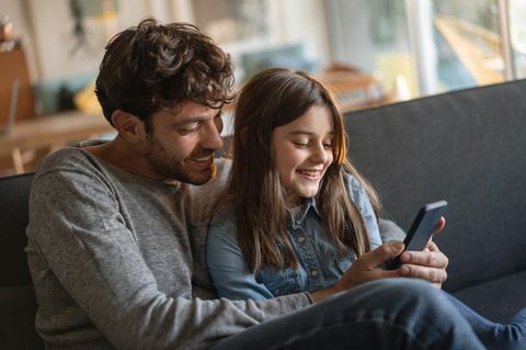 Handyvertrag für Kinder: Vater zeigt seiner Tochter auf der Couch ein Video auf dem Smartphone