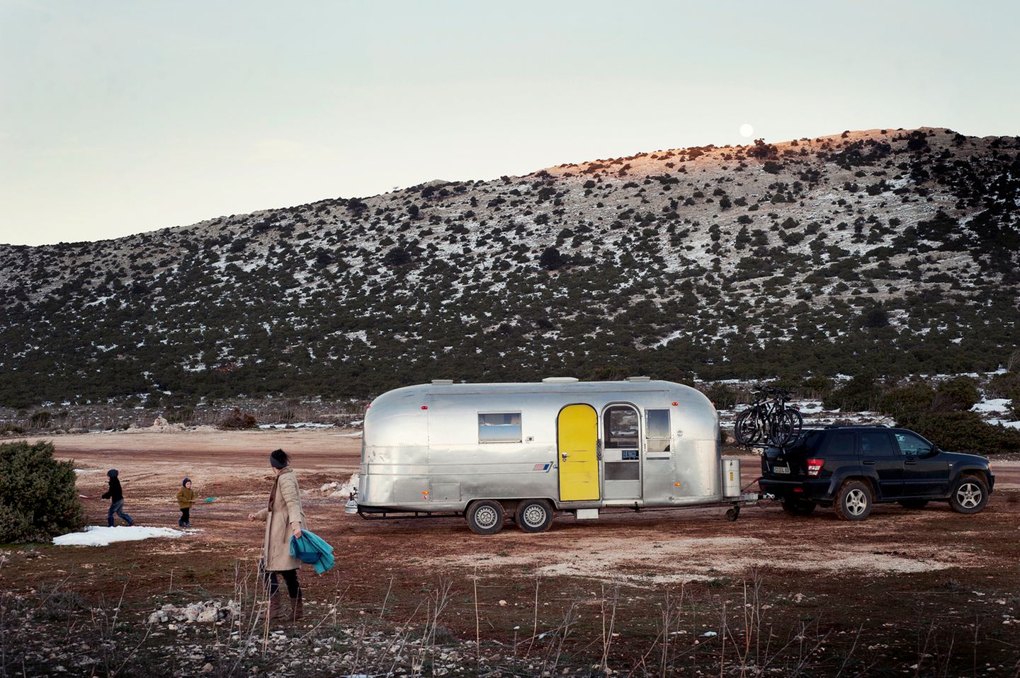 Vor dem geparkten Airstream spielen die beiden Jungs in den letzten Schneeresten. Im Hintergrund karge Berge.