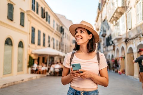 Junge Touristin wandert mit Sonnenhut und Smartphone durch eine belebte mediterrane Straße