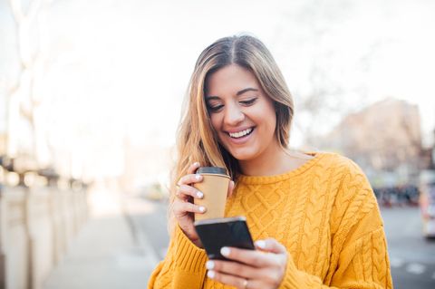 Junge Frau in gelbem Pullover mit Kaffee und Smartphone in der Hand