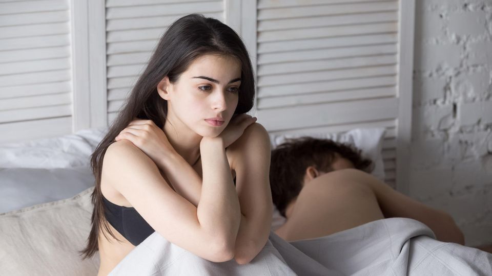 Kein Sex: Eine Frau sitzt frustriert im Ehebett neben ihrem schlafenden Partner