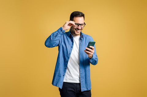 Mann mittleren Alters vor orangem Hintergrund schaut lachend auf sein Smartphone