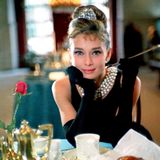 Audrey Hepburns sitzt im schwarzem Kleid mit einer Zigarette in der Hand an einem gedecktem Tisch