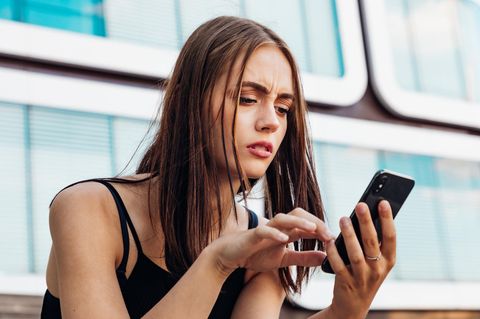 Junge Frau hält mit verwirrtem Gesichtsausdruck ihr Handy in der Hand