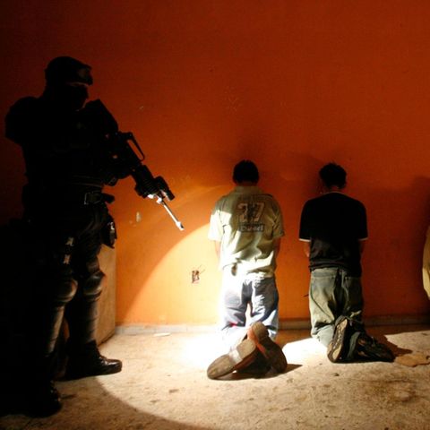 Gefangene knien vor einer Wand, dahinter ein bewaffneter Beamter