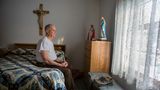 Ein alter Mann sitzt auf seinem akurat gemachtem Bett und schaut aus dem Fenster. Ein Kreuz und einem Marienstatue sind auch da.