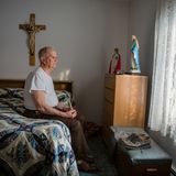 Ein alter Mann sitzt auf seinem akurat gemachtem Bett und schaut aus dem Fenster. Ein Kreuz und einem Marienstatue sind auch da.