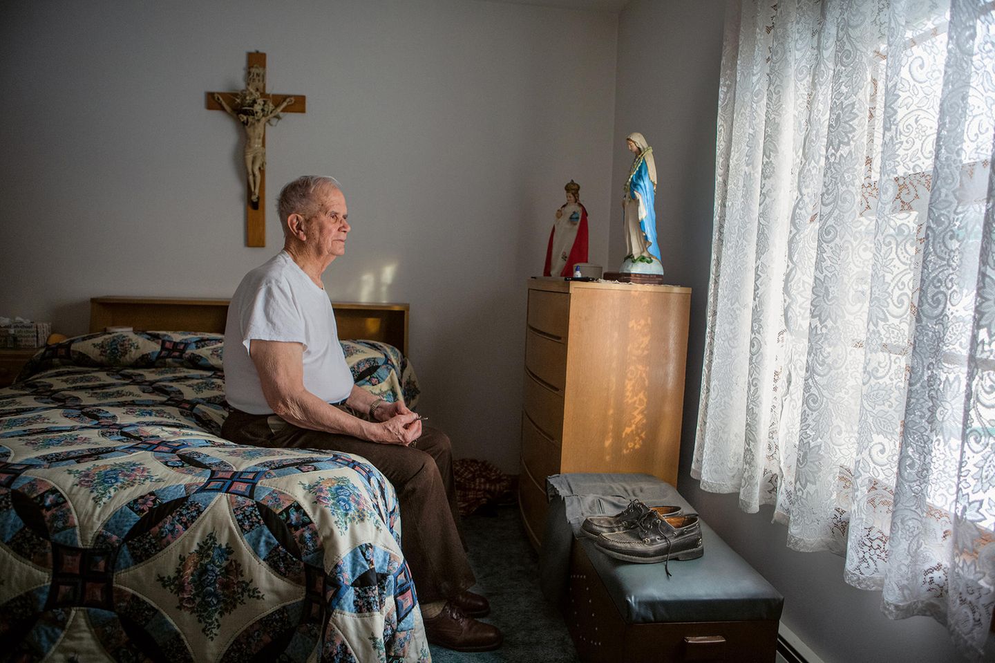 Ein alter Mann sitzt auf seinem akurat gemachtem Bett und schaut aus dem Fenster. Ein Kreuz und einem Marienstatue sind auch da. Ein alter Mann sitzt auf seinem akurat gemachtem Bett und schaut aus dem Fenster. Ein Kreuz und einem Marienstatue sind auch da.