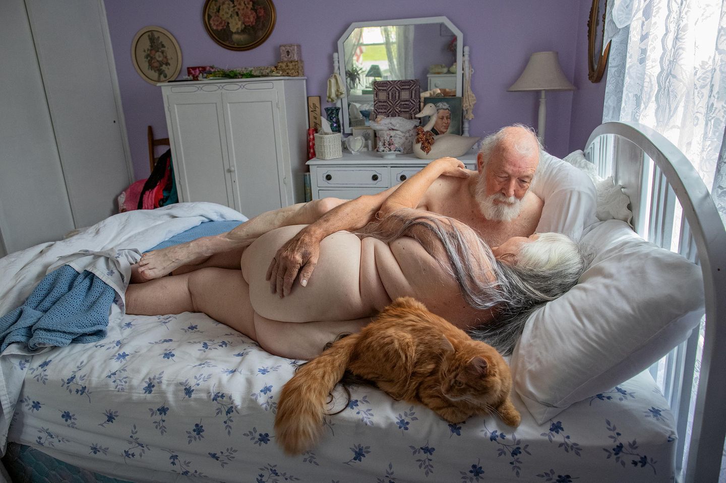 Ein altes Ehepaar liegt nackt auf dem Bett in einem weiß/lila Schlafzimmer und schaut sich an. Ein altes Ehepaar liegt nackt auf dem Bett in einem weiß/lila Schlafzimmer und schaut sich an.