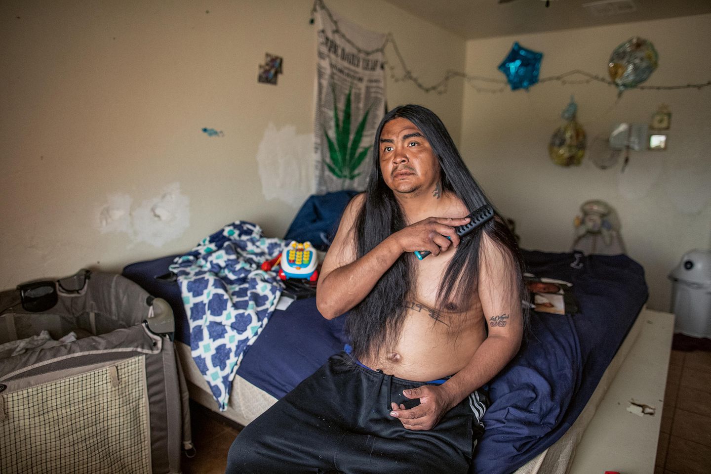 Ein Mann mit indigenem Hitnergrund sitzt auf seinem Bett und kämmpt sich mit einer Bürste durch seine langen, schwarzen Haare. Ein Mann mit indigenem Hitnergrund sitzt auf seinem Bett und kämmpt sich mit einer Bürste durch seine langen, schwarzen Haare.
