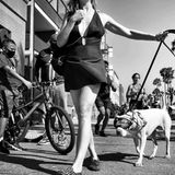 Frau im Kleid hält ihren Hund mit ausgestrecktem Arm an der Leine, welcher eine Skibrille und ein Bandana trägt