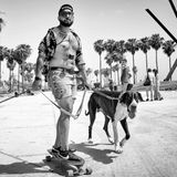 Ein rauchender Mann mit offenem Hemd fährt Longboard und hält zwei große Hunde an der Leine